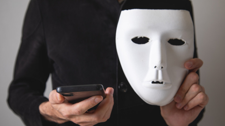 A  person wearing black with a white mask in one hand and an iPhone in the other.